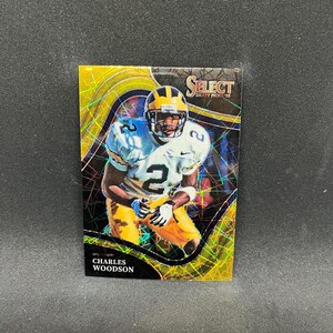 May include: A Charles Woodson football trading card from the Select Draft Picks '22 series. The card features a player in a white and gold uniform with the number 2. The background has a gold and iridescent design.