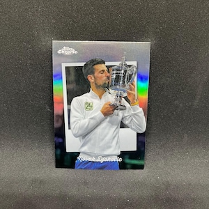 Puede incluir: Una tarjeta coleccionable Topps Chrome con Novak Djokovic besando un trofeo. Djokovic lleva una chaqueta blanca con un parche verde. La tarjeta tiene un fondo holográfico y el texto "Novak Djokovic".