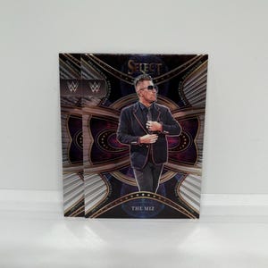 Puede incluir: Tres cromos WWE Select con The Miz. Las tarjetas tienen un fondo oscuro con detalles dorados y el logo de la WWE. The Miz lleva un traje negro y gafas de sol.
