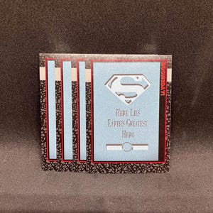 Puede incluir: Una tarjeta coleccionable con fondo azul claro, el logo de Superman y el texto "Here Lies Earth's Greatest Hero". La tarjeta tiene un borde negro y rojo con rayas azules verticales.