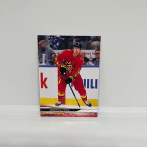 May include: A hockey trading card featuring Brady Tkachuk in a red and yellow uniform. The card shows Tkachuk in a skating pose, holding a hockey stick. The background includes a hockey rink and the text "BRADY TKACHUK" and "SENATORS".