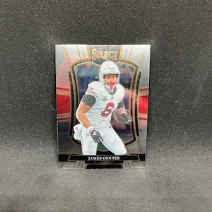 May include: A James Conner football trading card from the Select 2023 series. The card features a player in a white uniform with the number 6, holding a football. The card has a silver and red design with the text "JAMES CONNER" and "CARDINALS".