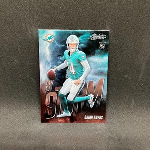 May include: A football trading card featuring Quinn Ewers in a teal and white uniform, holding a football. The card has the text "BY STORM" and "Absolute Football" with a Miami Dolphins logo and lightning background.