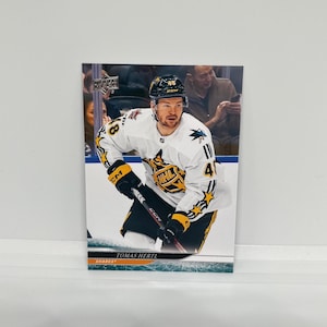 以下が含まれることがあります： サンノゼ・シャークスのトーマス・ハーテル選手が写ったホッケーカード。NHLのロゴと星のアクセントが入った白と黒のホッケーユニフォームを着ています。カードにはUpper Deckのロゴが入っています。