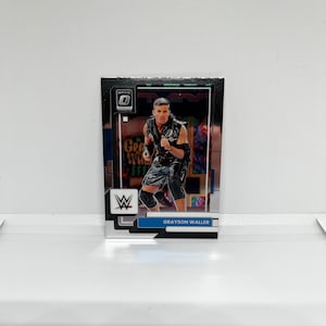 Puede incluir: Una tarjeta de intercambio con un luchador con un atuendo blanco y negro. La tarjeta tiene un fondo plateado con un borde azul y blanco. La tarjeta tiene el texto "OPTIC" y "GRAYSON WALLER".
