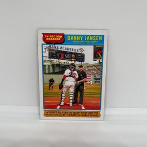 May include: A baseball card featuring Danny Jansen of the Boston Red Sox. The card has a red and blue color scheme with text that reads "24 Record Breaker" and "First Player in MLB History to Play on Both Teams in Same Game."