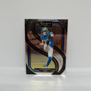 May include: A trading card depicting a football player in a blue and yellow uniform kicking a ball. The card has a black and gold border with the word "SELECT" at the top. The player's name, Joshua Karty, and the team name, RAMS, are printed at the bottom.
