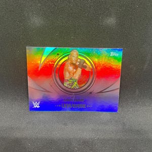 May include: A Topps trading card with a holographic design, featuring a wrestler. The card has a central image of the wrestler, surrounded by a circular design. The card includes the WWE logo and the text "FOR... UNIVERSE..."