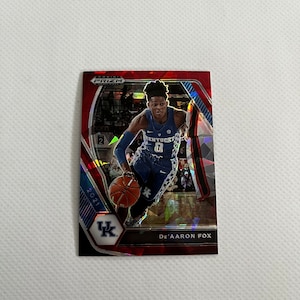 May include: A red, white and blue basketball trading card featuring a basketball player wearing a blue jersey with the number 0 and the word "Kentucky" on it. The card is from the 2021 Panini Prizm Basketball set and features De'Aaron Fox.