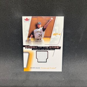 May include: A Fleer Genuine trading card featuring Brian Giles of the Pittsburgh Pirates. The card displays a photograph of the player in a white uniform, the text Names of the Game, and an embedded piece of white fabric from authentic game-worn pants.