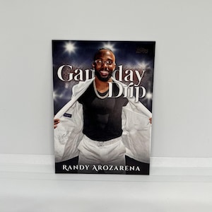 May include: A trading card featuring Randy Arozarena. The card has the words "Gameday Drip" in white and black text. Arozarena is wearing a white suit jacket over a black shirt and gold chain. The background has a dark, starry design.