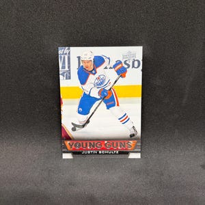 May include: A rectangular Upper Deck hockey trading card featuring Justin Schultz in an Edmonton Oilers uniform. The card displays the text Young Guns and Justin Schultz at the bottom, with the player holding a hockey stick on the ice.