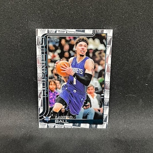 Puede incluir: Una tarjeta de baloncesto con LaMelo Ball con un uniforme púrpura de los Hornets, sosteniendo un balón de baloncesto. La tarjeta tiene un borde plateado con un patrón de red de baloncesto y el logotipo de Topps. El nombre del jugador está impreso en la parte inferior.