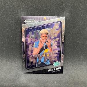 Puede incluir: Una tarjeta coleccionable Prizm Draft Picks con Andrew Marsh con uniforme azul y amarillo. La tarjeta tiene un fondo holográfico plateado con la palabra "HYPE" en letras grandes y estilizadas. La tarjeta también incluye el texto "Rated Prospect" y "Wolverines."