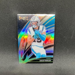 May include: A football trading card featuring Adam Thielen of the Carolina Panthers. The card has a holographic design with blue and silver accents. Thielen is wearing a white and blue uniform with the number 23.