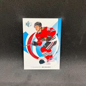 Puede incluir: Una tarjeta de hockey con Connor Bedard en un uniforme rojo y negro de los Blackhawks de Chicago. La tarjeta tiene un diseño azul y blanco con el logotipo SP en la parte superior. El jugador sostiene un palo de hockey.