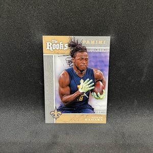 Peut inclure: Carte de collection représentant Alvin Kamara en uniforme des Saints de la Nouvelle-Orléans. La carte a une bordure dorée et argentée avec le texte "The Rookies" et "Panini". Kamara tient un ballon de football, portant un maillot bleu et des gants verts.