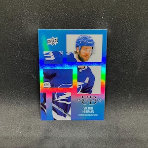 Op de afbeelding: Een hockey trading card met Victor Hedman van de Tampa Bay Lightning. De kaart heeft een blauw en holografisch ontwerp met de Upper Deck en UD Portraits logo's. Hedman draagt een blauw shirt en helm.
