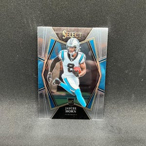 May include: A 2021 Select RC Jaycee Horn Panthers football trading card. The card features a player in a Carolina Panthers uniform, running with a football. The card has a silver and blue design.