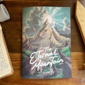 May include: A book titled "The Eternal Mountain" with a mountain landscape illustration on the cover. The cover art features a mountain range, sky, and the title in white script. The book is on a wooden surface with other objects.
