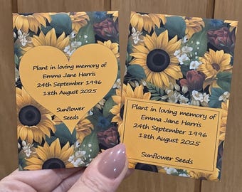 May include: Two packets of sunflower seeds with a floral design featuring yellow sunflowers and green leaves. Each packet has a yellow label with text that reads "Plant in loving memory of Emma Jane Harris 24th September 1996 18th August 2025 Sunflower Seeds."