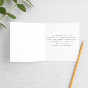 Open white greeting card mock-up showing printed message inside with pencil and green leaves on white wooden background. Font sample displayed for optional personalised message insert.