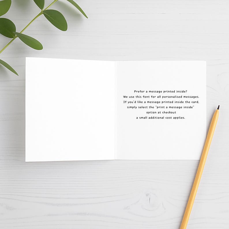 Open white greeting card mock-up showing printed message inside with pencil and green leaves on white wooden background. Font sample displayed for optional personalised message insert.