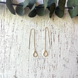May include: A pair of gold teardrop earrings hanging from delicate silver chains against a white wooden background.