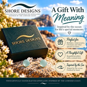 Puede incluir: Un par de pendientes azul claro con postes de plata marcados 925, colocados frente a un joyero verde oscuro con el texto dorado SHORE DESIGNS. El fondo muestra una costa con el texto A Gift With Meaning.