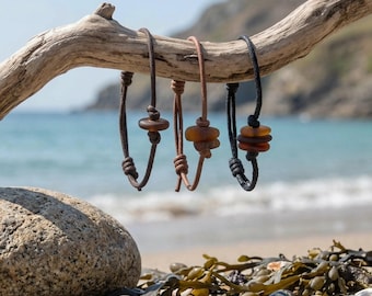 Puede incluir: Tres pulseras hechas de cordón fino en colores marrón oscuro, tostado y negro. Cada pulsera presenta una o varias piezas de vidrio marino de color ámbar, lisas y redondeadas, aseguradas mediante nudos a lo largo del cordón ajustable.
