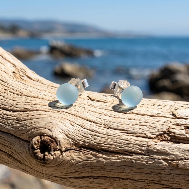 Puede incluir: Un par de pendientes de bot&oacute;n con cuentas redondas de vidrio marino esmerilado en color azul claro. Cada cuenta est&aacute; montada sobre un poste de plata pulida con un sello 925 visible, creando un dise&ntilde;o sencillo para uso diario.