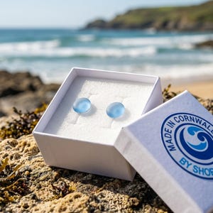 Puede incluir: Un par de pendientes circulares peque&ntilde;os, lisos y de color azul claro dentro de una caja de regalo cuadrada blanca. La tapa abierta presenta un logotipo azul circular con el texto Made in Cornwall by Shore impreso en la esquina exterior.