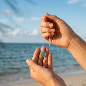 May include: A silver necklace featuring a seafoam green, teardrop-shaped pendant with a heart-shaped cutout. The necklace is held up against a backdrop of the ocean and sky.