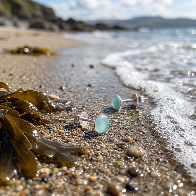 Puede incluir: Un par de pendientes de bot&oacute;n con piedras cabuj&oacute;n redondas y transl&uacute;cidas de color azul claro, montadas en postes de metal plateado. Los pendientes est&aacute;n colocados sobre una superficie arenosa cerca del agua, con algas marrones cerca.