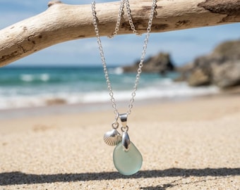 May include: A silver chain necklace featuring two pendants: a small, textured silver seashell charm and a smooth, translucent, pale seafoam green piece of sea glass, both suspended from a polished silver bail on a delicate metal link chain.