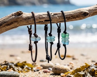 Puede incluir: Tres pulseras ajustables de cordón marrón oscuro y negro, cada una con cuentas de vidrio marino pulido. La primera pulsera tiene una cuenta azul claro, la segunda tiene dos cuentas azul claro y la tercera tiene tres cuentas azules y verdes.