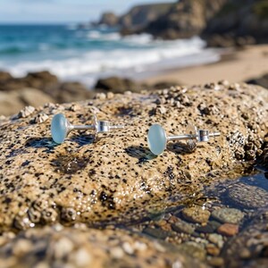 Puede incluir: Un par de pendientes de bot&oacute;n con cabujones de vidrio marino azul claro, redondos y lisos, montados en postes de metal plateado pulido. Los pendientes est&aacute;n colocados sobre una superficie de roca texturizada y moteada.
