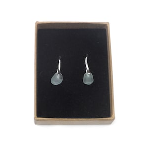 May include: A pair of earrings with silver hooks and pale blue, teardrop-shaped stones. The earrings are presented in a small, open, brown cardboard box with a black interior. The background is white.