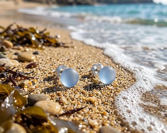 Puede incluir: Un par de pendientes de botón con cabujones de vidrio azul claro, redondos y translúcidos, montados en postes de metal plateado. Los pendientes están colocados sobre una superficie de arena de playa con guijarros y algas.