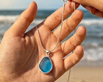 Puede incluir: Un collar de plata con un colgante de piedra azul de forma irregular. El colgante está engastado en un bisel de plata y cuelga de una delicada cadena de plata. El fondo muestra una playa y el mar.