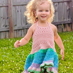 May include: A young girl wearing a pink, green, and blue crochet dress with a ruffled hem. She is standing in a grassy field and smiling.