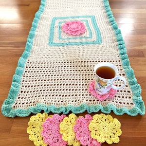 Könnte beinhalten: Ein gehäkelter Tischläufer mit einem hellgrünen Rand und einer rosa Blume in der Mitte. Vier gehäkelte Untersetzer in Gelb und Rosa liegen auf einer Holzoberfläche.