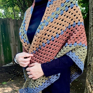 May include: A crocheted shawl with a multi-colored striped pattern in shades of brown, green, and blue. The shawl is made with a granny square stitch and has a fringed edge. @SimpleThingsCrochet