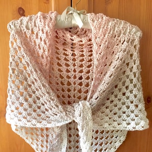 Puede incluir: Un chal de ganchillo en un degradado de rosa y blanco. El chal está atado en un nudo y colgado de una percha de madera. El texto "@SimpleThingsCrochet" es visible en la esquina inferior derecha de la imagen.