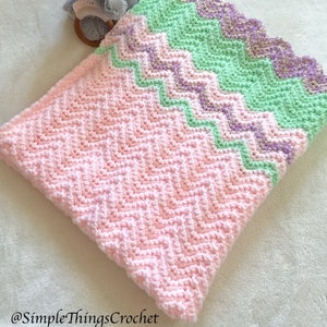 May include: A pink, green, and purple chevron crochet baby blanket with a scalloped edge. The blanket is lying on a white fuzzy surface with a gray elephant plush toy on top.