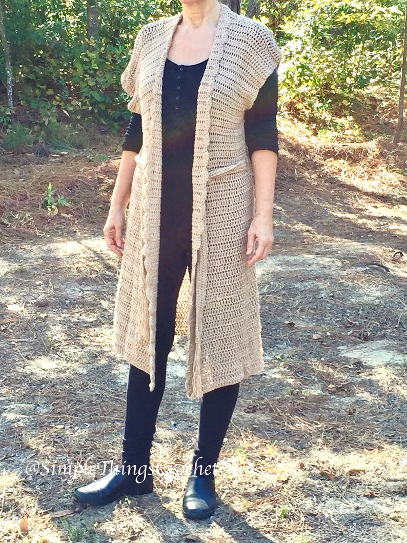Puede incluir: Un c&aacute;rdigan largo de crochet beige con cuello chal y detalles de flecos. El c&aacute;rdigan se lleva sobre una camiseta negra y leggings negros.