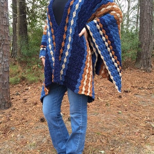 Puede incluir: Un chal de ganchillo hecho a mano, azul, marr&oacute;n y crema, con un dise&ntilde;o de cuello en V. El chal presenta un patr&oacute;n de rayas y se usa sobre un cuello alto negro y jeans azules. La imagen est&aacute; tomada al aire libre con &aacute;rboles en el fondo.