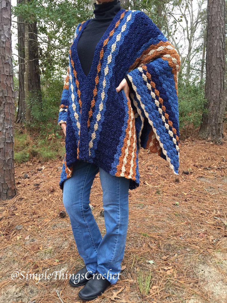 Puede incluir: Un chal de ganchillo hecho a mano, azul, marr&oacute;n y crema, con un dise&ntilde;o de cuello en V. El chal presenta un patr&oacute;n de rayas y se usa sobre un cuello alto negro y jeans azules. La imagen est&aacute; tomada al aire libre con &aacute;rboles en el fondo.