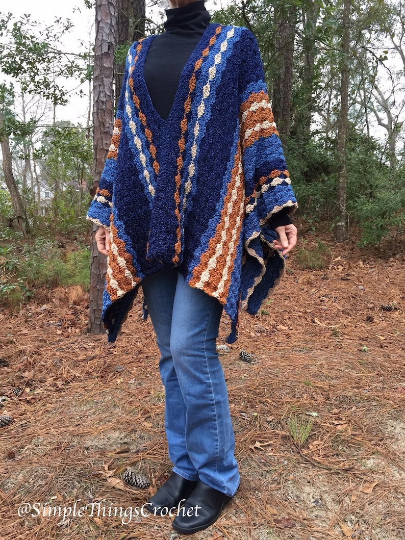 Puede incluir: Un poncho de ganchillo azul, marr&oacute;n y crema con cuello en V. El poncho presenta un patr&oacute;n geom&eacute;trico y se lleva sobre un cuello alto negro y vaqueros azules. La imagen est&aacute; tomada al aire libre.