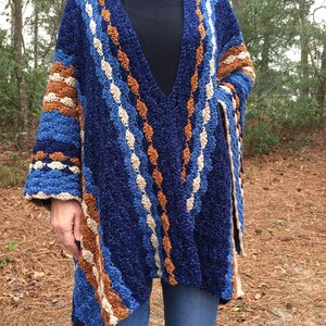 Puede incluir: Un chal de ganchillo hecho a mano, azul, marr&oacute;n y blanco, con cuello en V. El chal presenta un patr&oacute;n texturizado con rayas de varios colores y un ajuste holgado. Las mangas son anchas y el chal se lleva sobre un cuello alto negro y vaqueros azules.